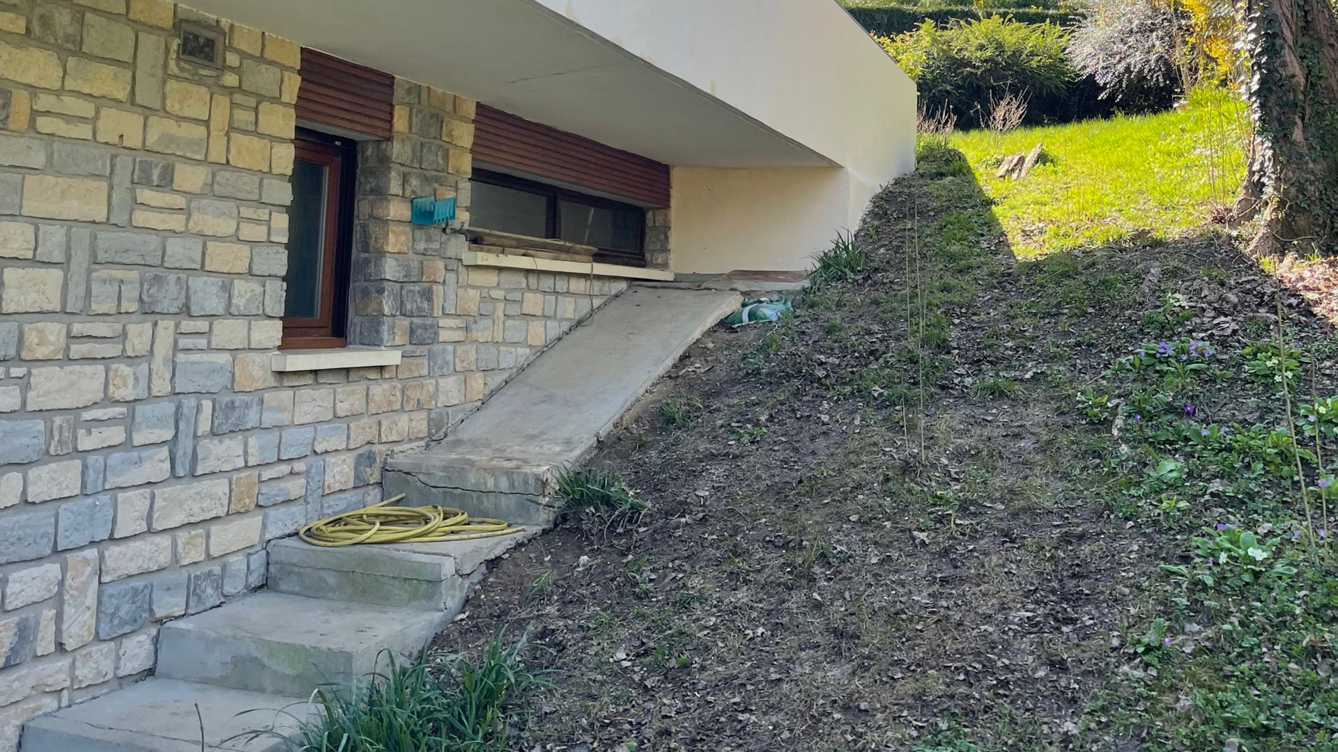 Vue extérieure d'un bâtiment avec mur en pierre, rampe en béton, marches et flanc de colline envahi par la végétation.