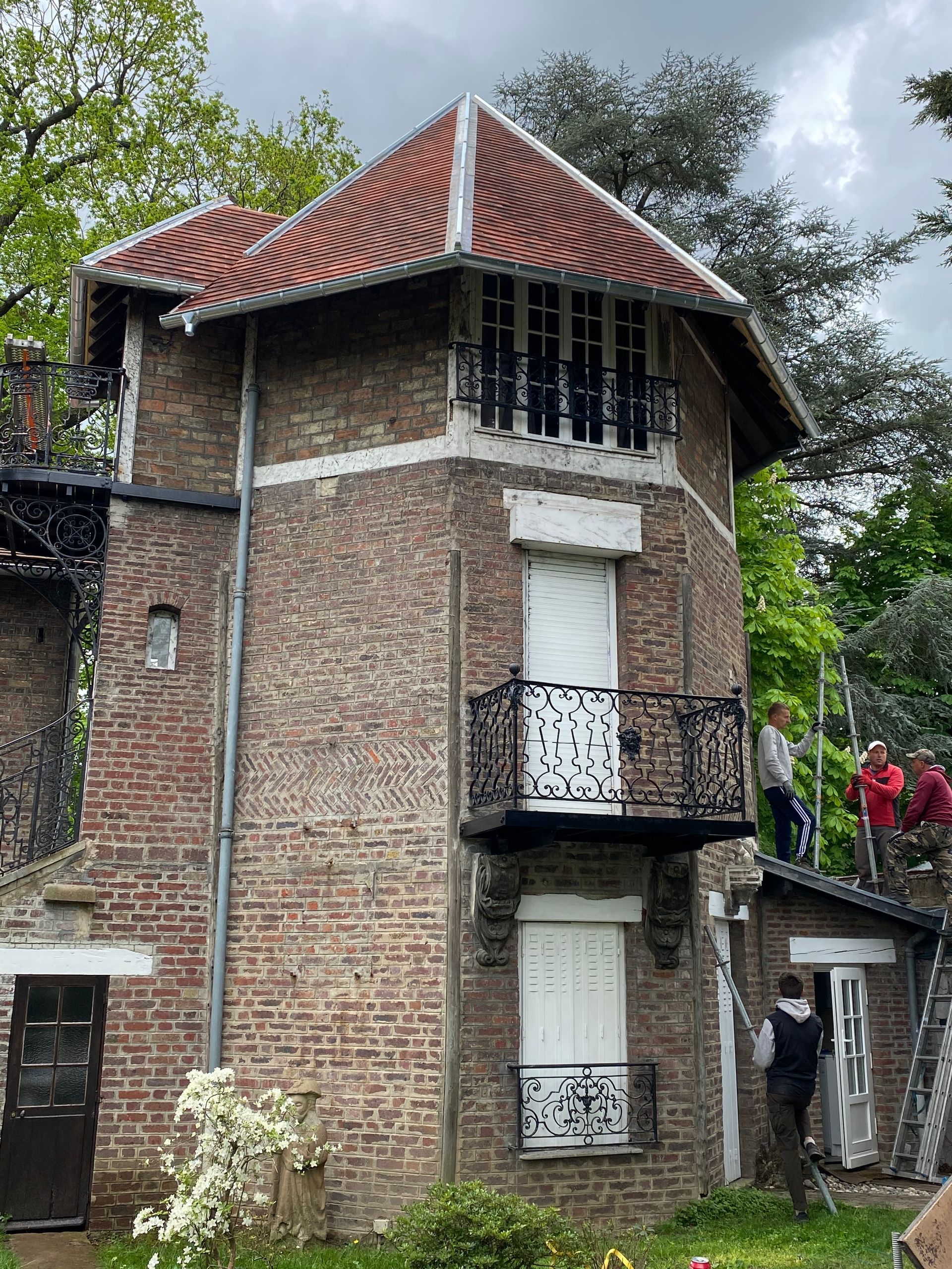 Bâtiment en briques avec une tour octogonale et des balcons en fer forgé. Des personnes travaillent sur le toit.