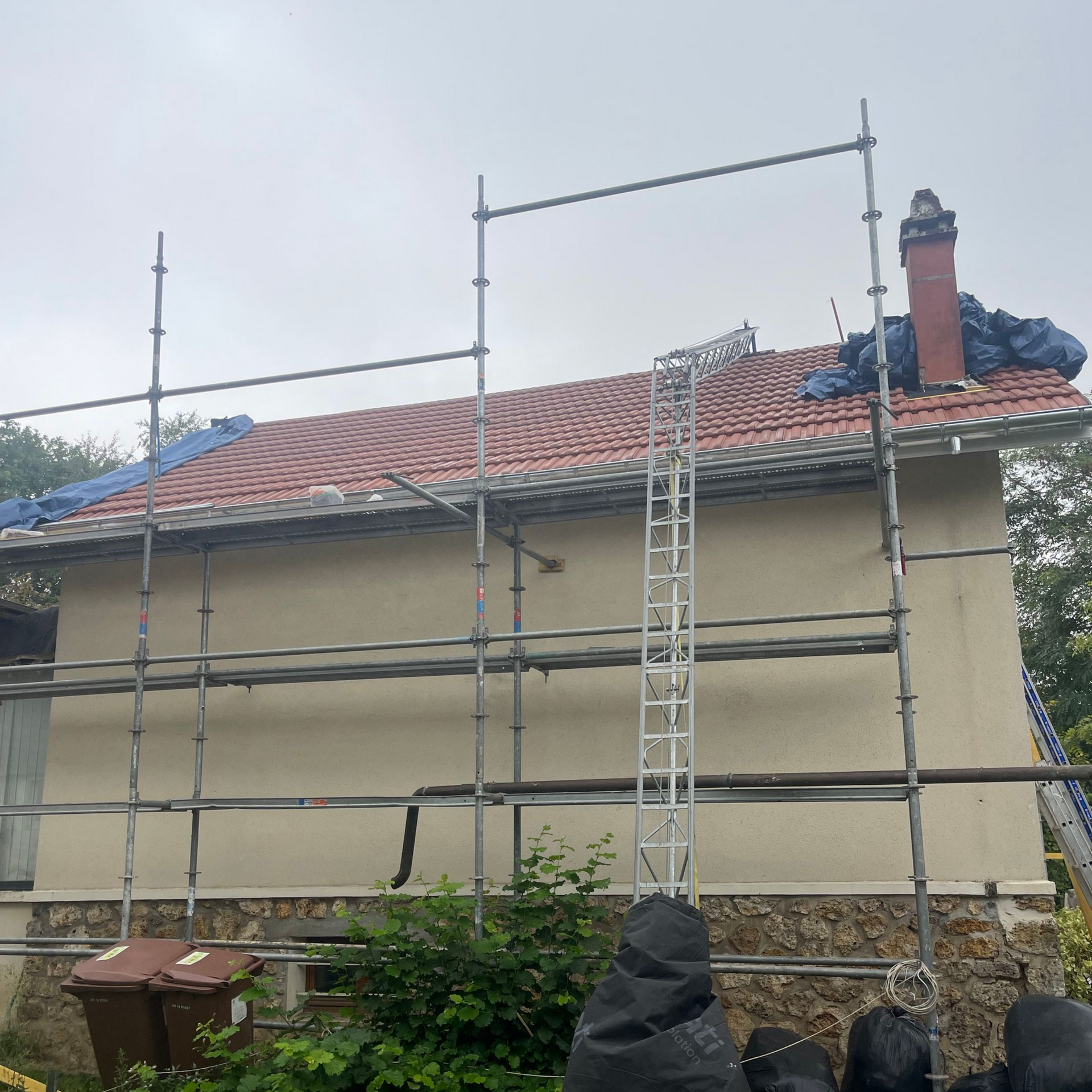 Des échafaudages entourent une maison au toit de tuiles rouges, avec une cheminée et des murs en stuc beige, sous un ciel nuageux.