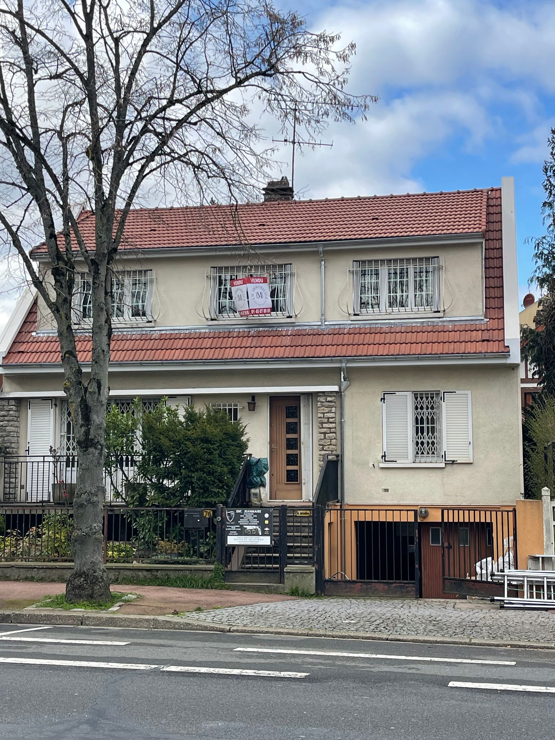 Maison à deux étages avec toit de tuiles rouges, murs beiges et fondations partiellement apparentes par temps nuageux.