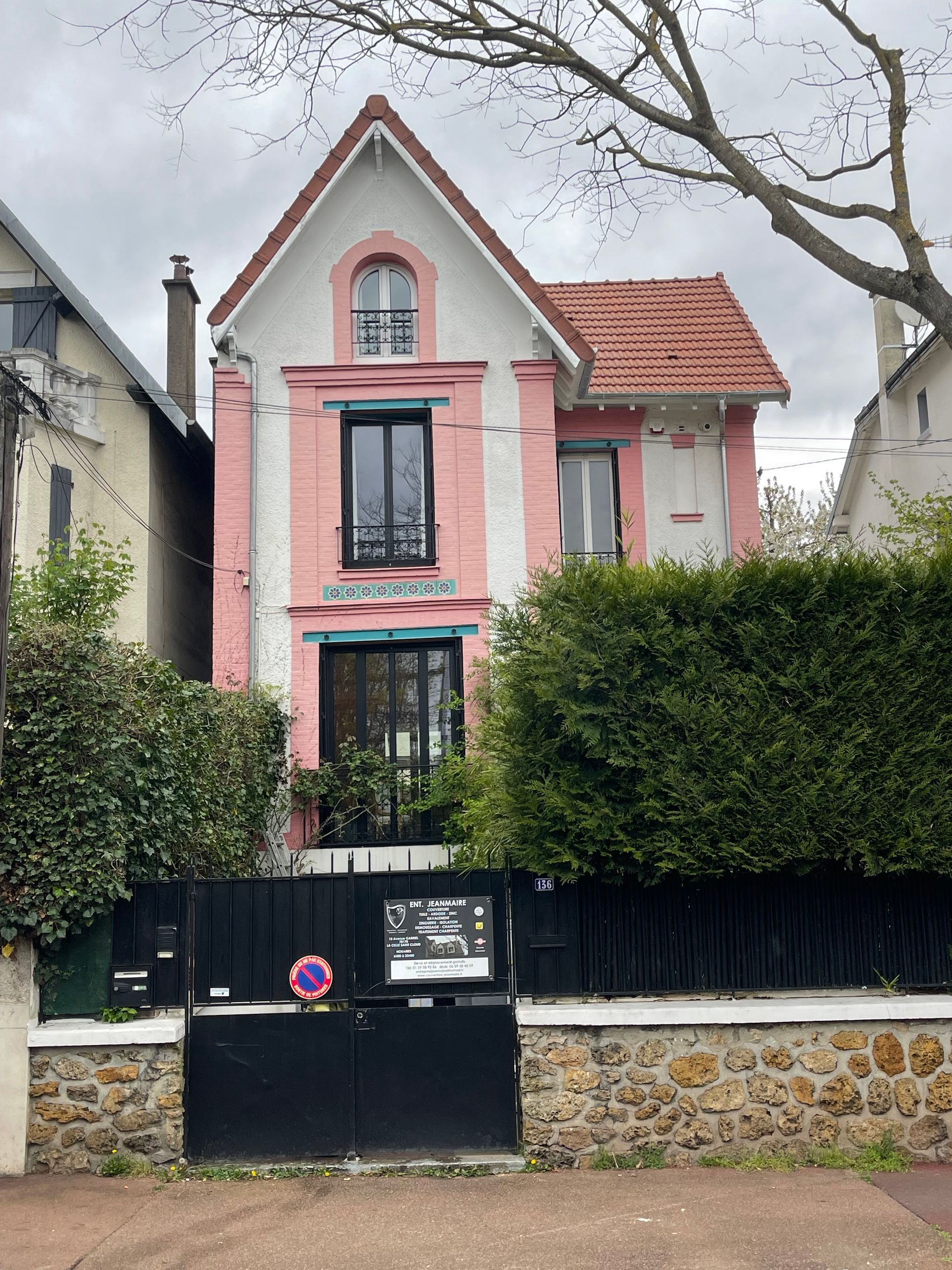 Maison rose et blanche avec une clôture noire et un toit rouge sous un ciel nuageux.