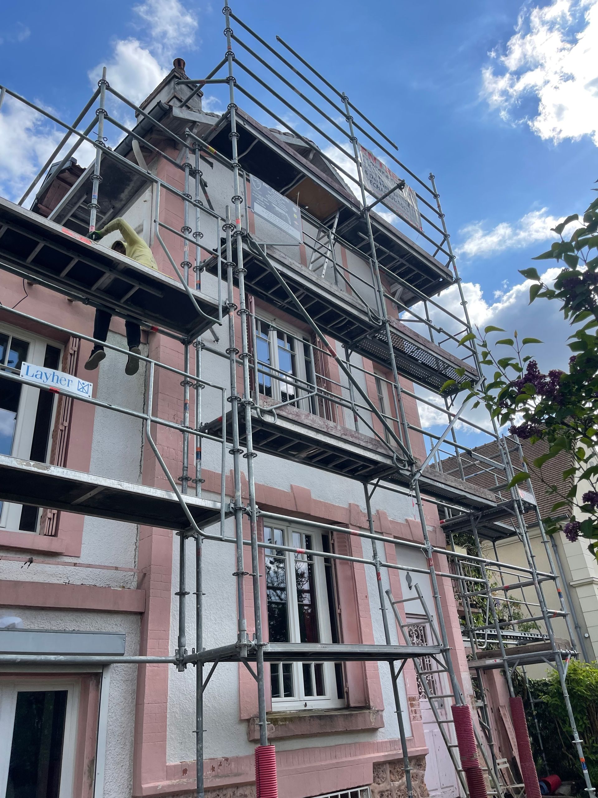 Des échafaudages entourent un bâtiment rose et blanc en rénovation, sur fond de ciel nuageux.