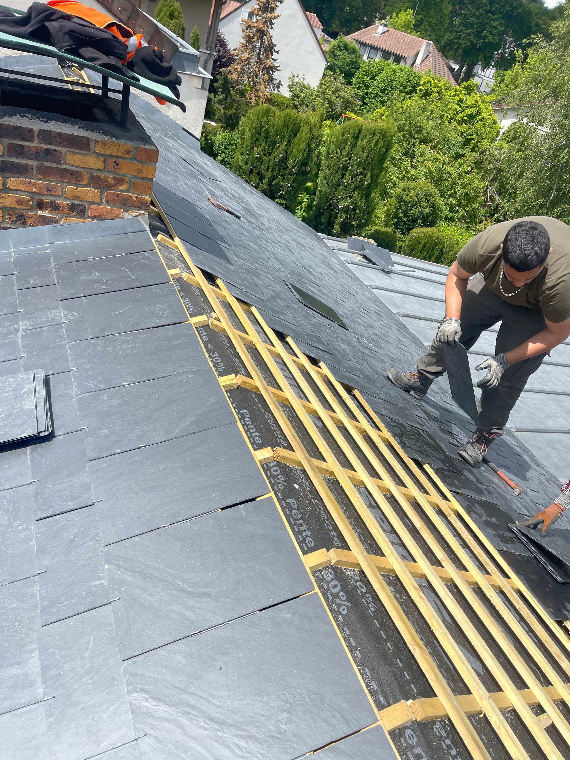 Couvreur sur un toit, posant des ardoises. Sous-couche de toiture noire, poutres en bois, arbres verts en arrière-plan.