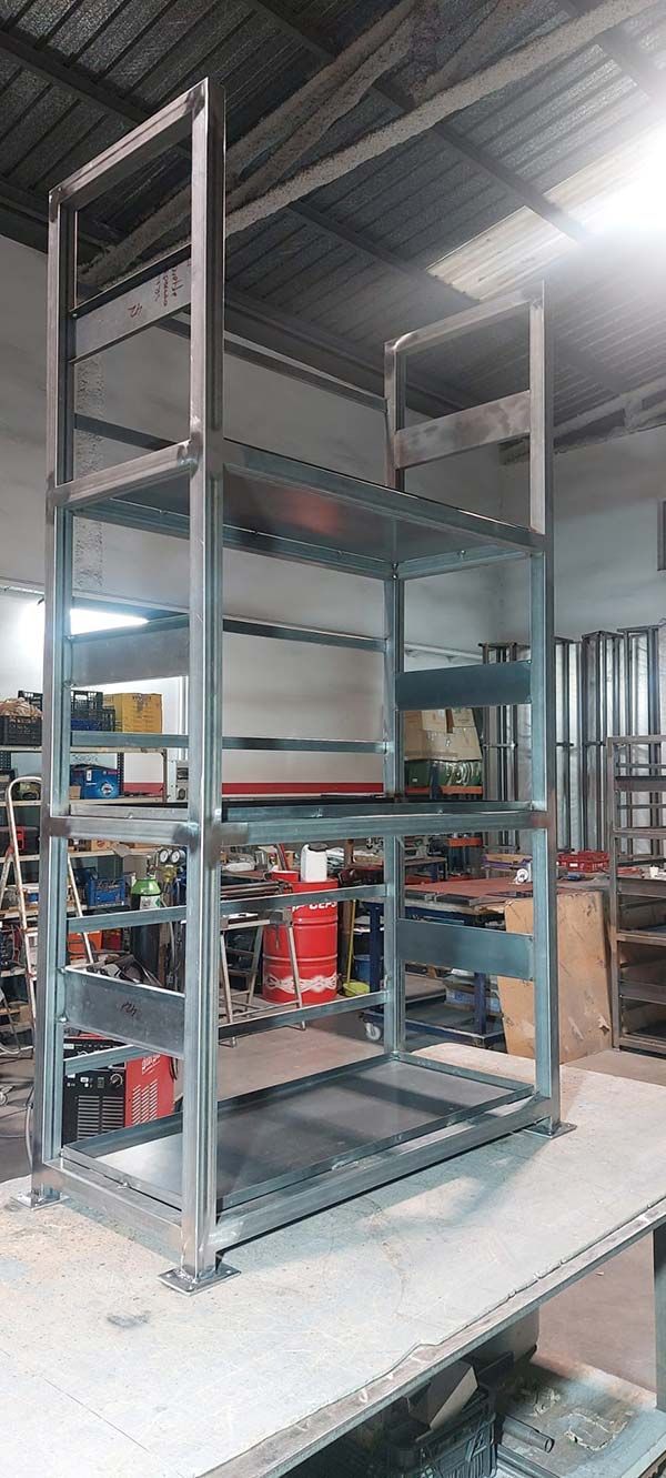Estantería alta de metal gris en un taller o almacén, colocada sobre un banco de trabajo con herramientas al fondo.