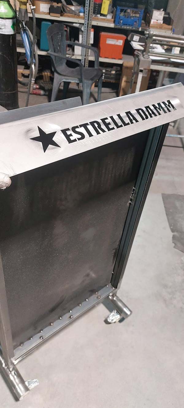 Soporte metálico de la marca Estrella Damm con panel frontal negro en un entorno de taller.