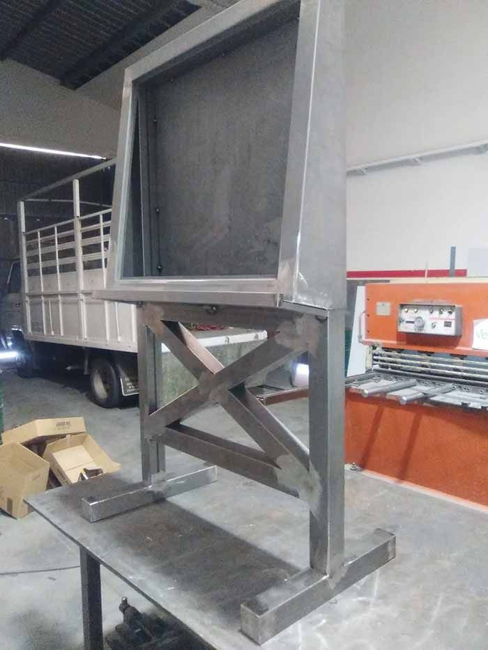 Gran estructura metálica gris para maquinaria con refuerzos transversales, mostrada en un almacén junto a un camión y una barrera de seguridad naranja.