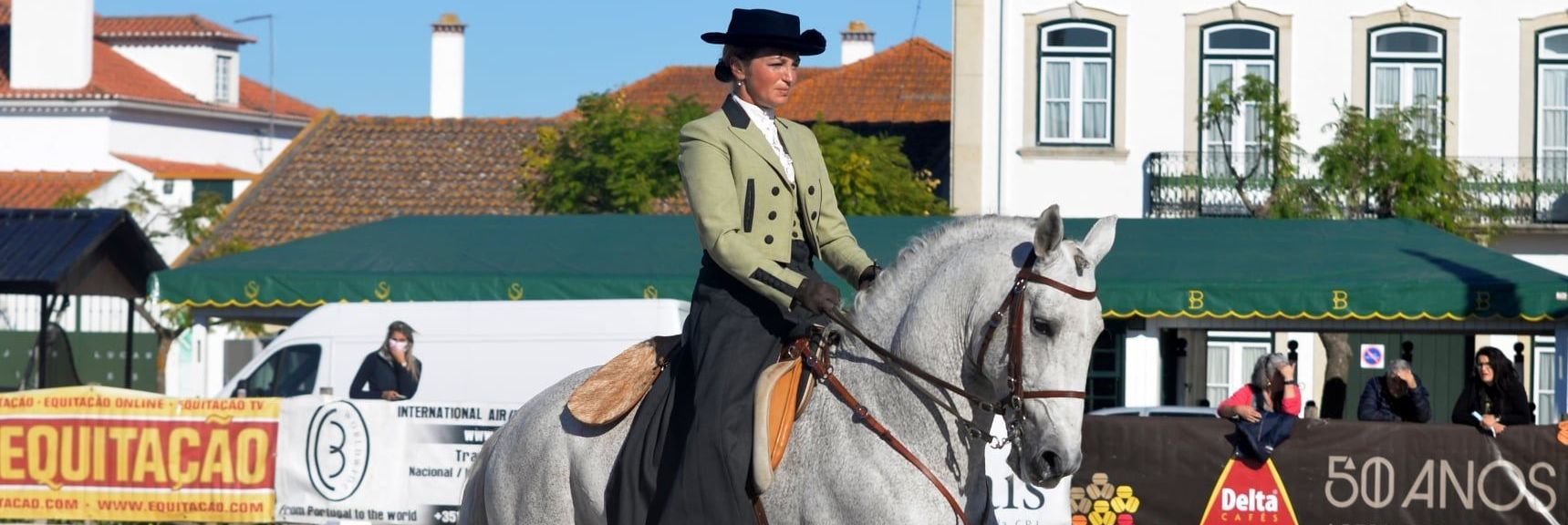 A woman is riding a horse in front of a sign that says equitatacao