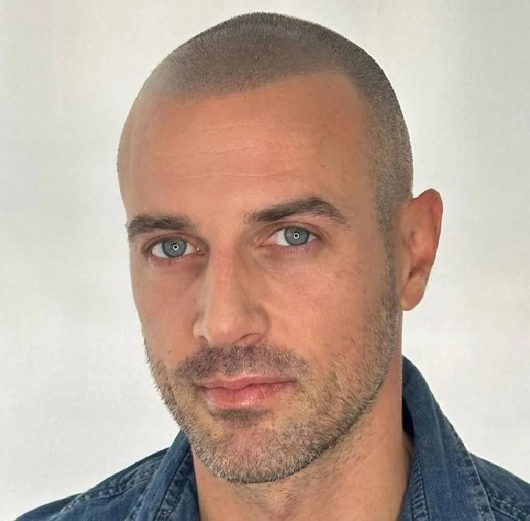 Hombre de pelo corto y ojos azules, vestido con camisa vaquera, mira hacia delante con expresión neutra.