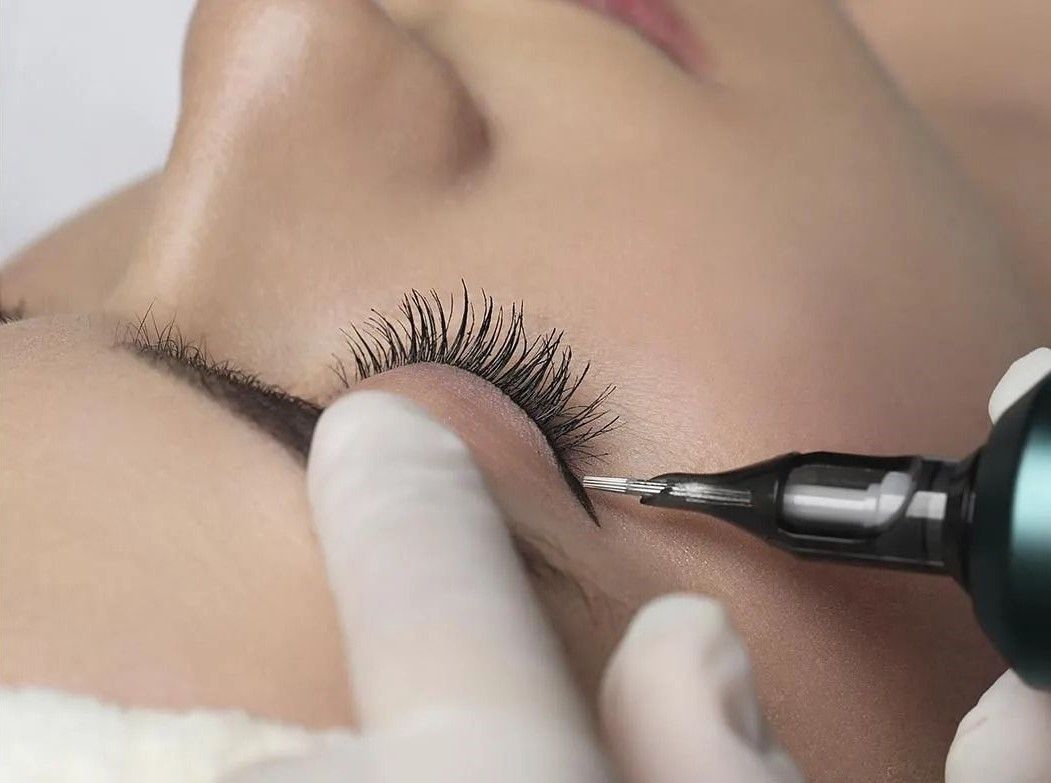 Persona que recibe un tatuaje de delineador de ojos, una mano enguantada sostiene el párpado, el lápiz de tatuaje aplica tinta en la línea de las pestañas.