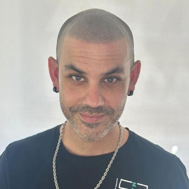 Hombre con cabeza rapada, camisa negra, collar de cadena y medidores sonriendo a la cámara.