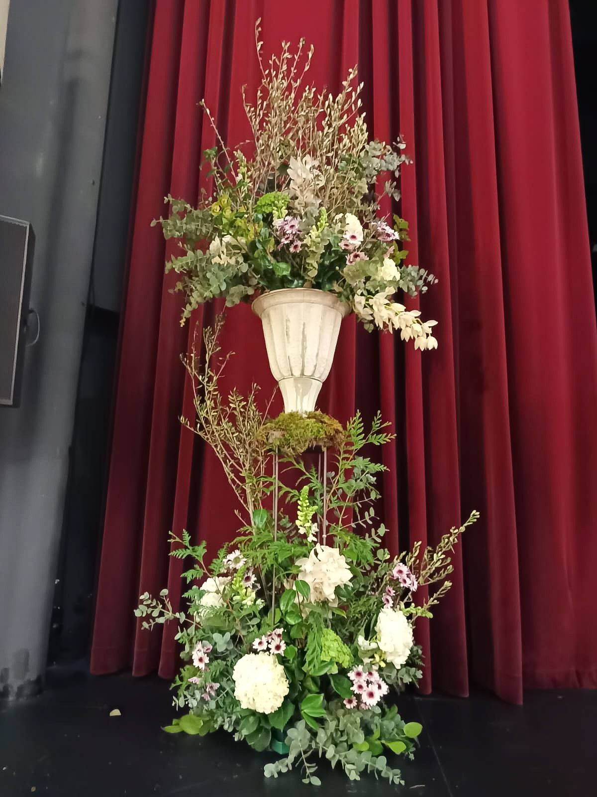 Un jarrón lleno de flores está sentado en un escenario frente a una cortina roja.