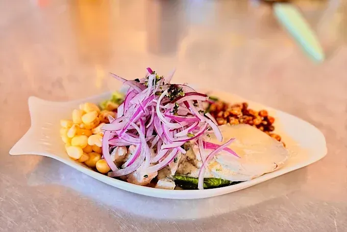 Ceviche en plato en forma de pescado, cubierto con cebolla morada y cilantro, con maíz y granos de maíz crujientes.