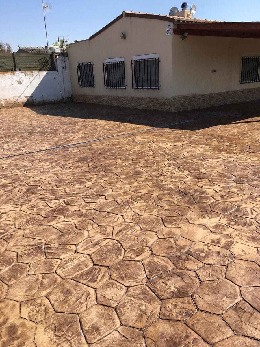 Vista exterior de una casa con pavimento estampado marrón; tres ventanas con contraventanas cerradas y una pared blanca.