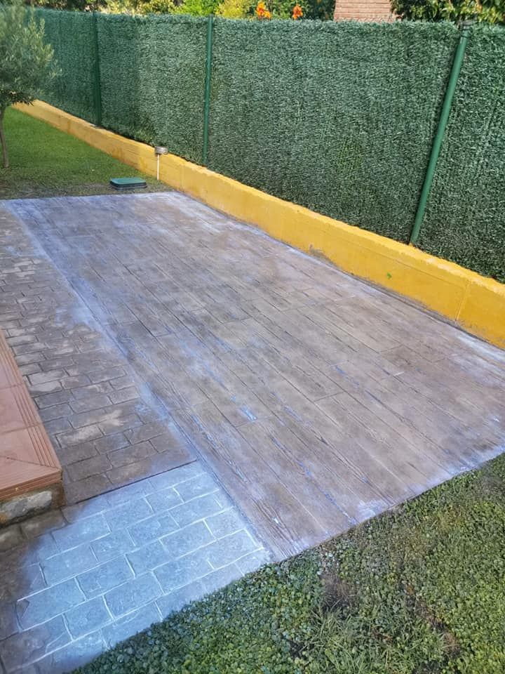 Patio de madera bordeado por un seto artificial amarillo de fondo y césped verde.
