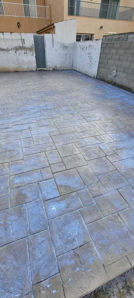 Zona pavimentada con bloques cuadrados de colores claros y muros circundantes. Al fondo, se ven edificios con balcones.