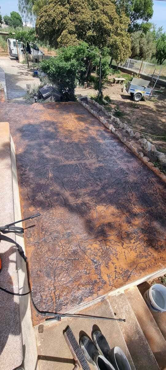Vista aérea de una superficie rectangular cubierta de material moteado oscuro, posiblemente alquitrán o asfalto, cerca de árboles y un edificio.