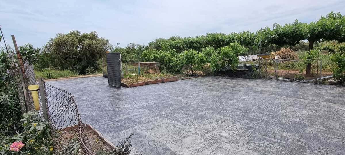 Zona de hormigón con muro de piedra y vegetación al fondo bajo un cielo nublado.