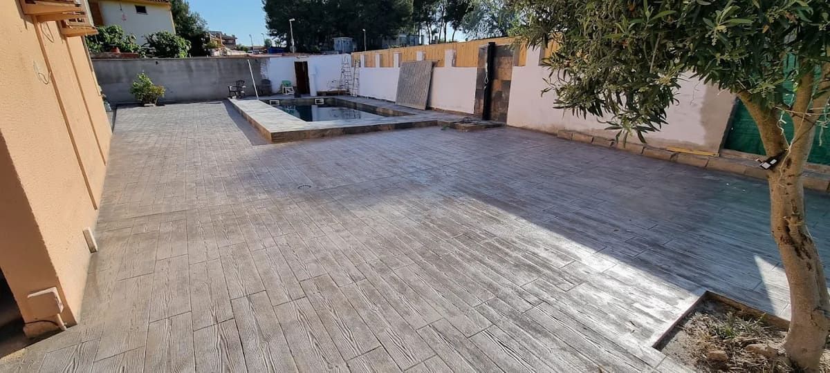 Un gran patio pavimentado con una piscina rectangular, rodeado de paredes blancas y un árbol.