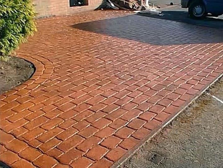Camino de entrada de hormigón con diseño de ladrillo, borde curvo, color marrón rojizo, al lado de un arbusto verde y un coche estacionado.