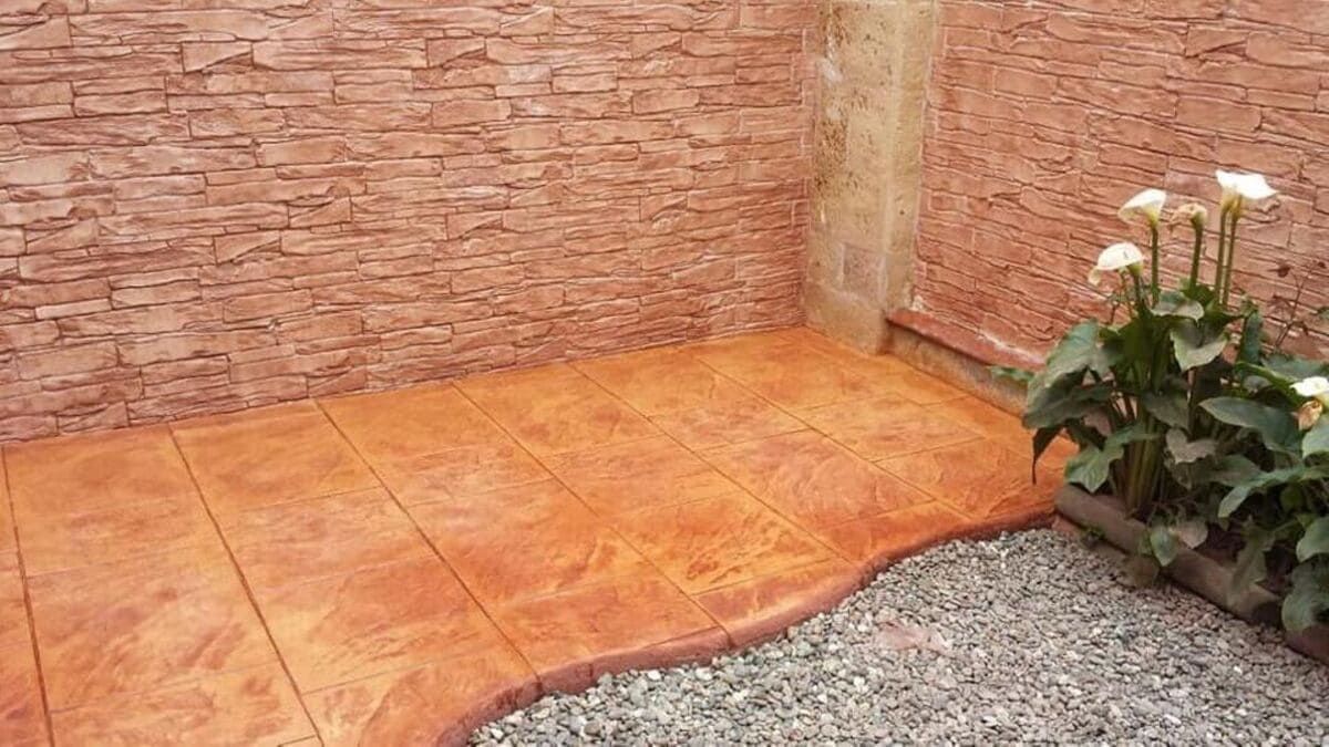 Patio exterior con pared tipo ladrillo, suelo de hormigón teñido, grava y calas en macetas; tonos marrón rojizos.