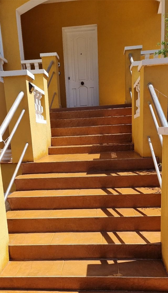 Entrada amarilla al edificio con escaleras que conducen a una puerta blanca. Barandillas blancas a ambos lados.