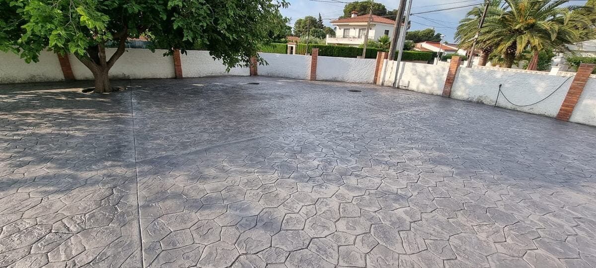 Un patio pavimentado rodeado de una pared blanca y un árbol. Se ve una casa a lo lejos.