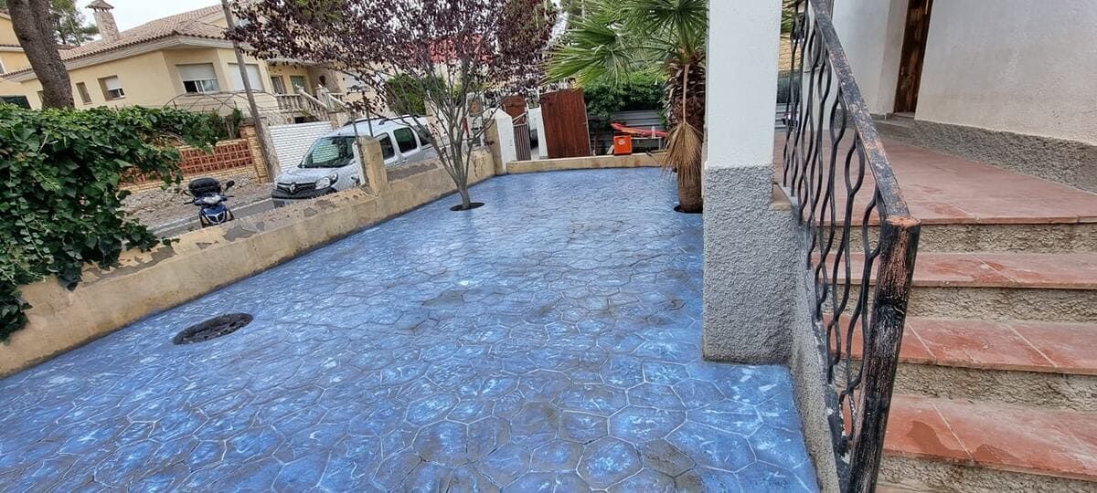 Entrada de hormigón pintado de azul con una casa, un vehículo, árboles y escaleras con barandilla decorativa.