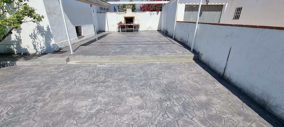 Un patio con piso pavimentado, paredes blancas y una parrilla para barbacoa en el fondo.