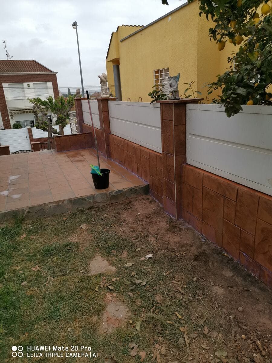 Exterior de una casa con pared de azulejos y jardín. Un cubo negro reposa sobre una superficie pavimentada.