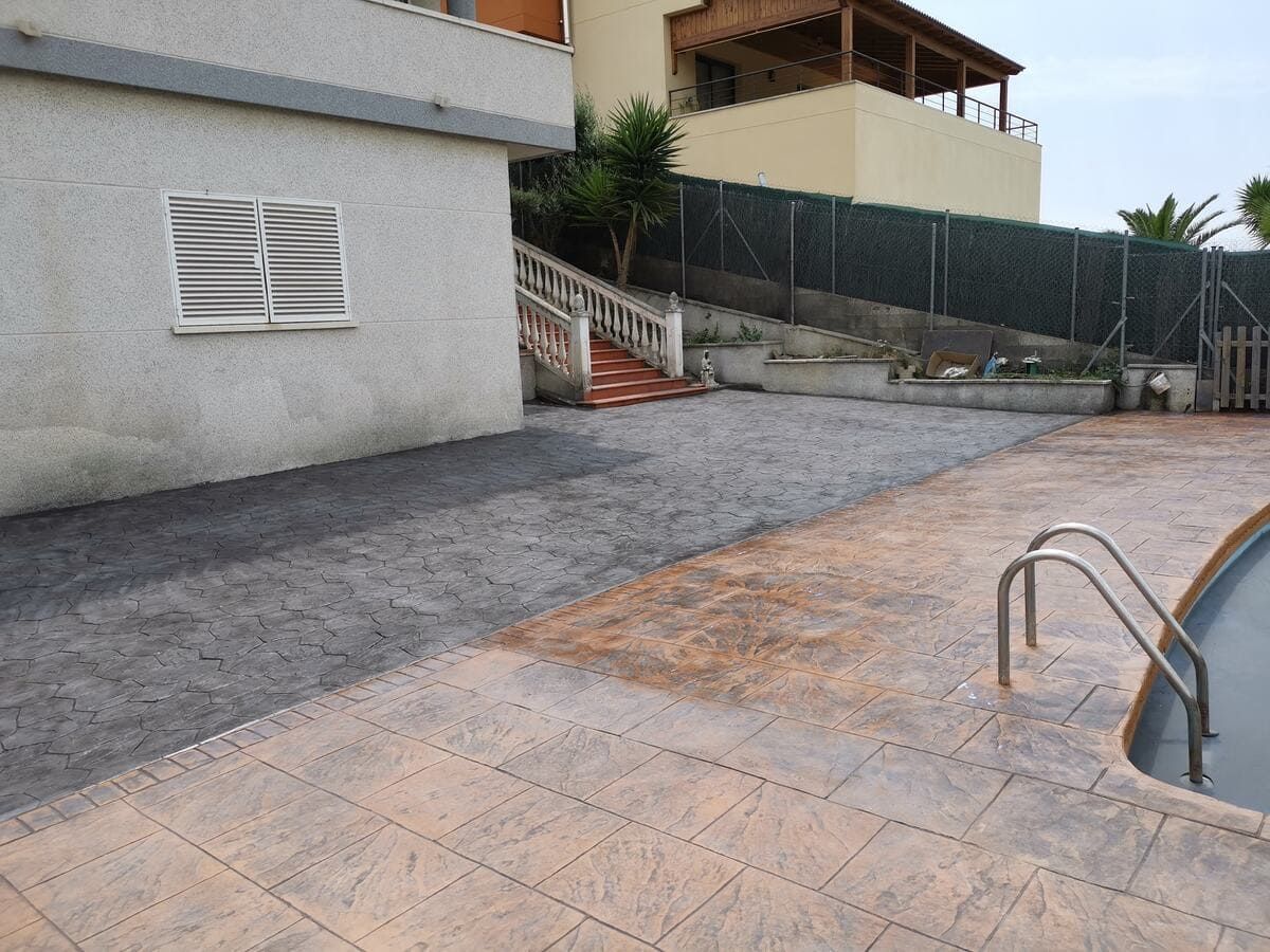 Patio de hormigón rodea una piscina junto a una casa de color claro.