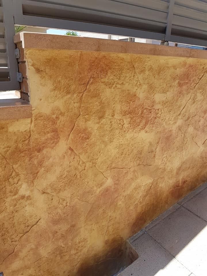 Muro de hormigón texturizado de color amarillo y marrón rojizo, con grietas visibles, situado bajo una estructura metálica, al lado de un escalón.