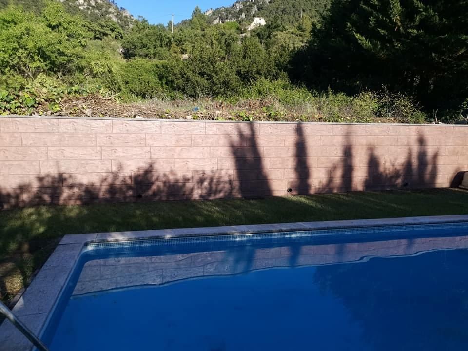 Una piscina rectangular refleja el cielo. Las sombras de los árboles se proyectan sobre una pared de ladrillo y el césped. Al fondo se ven montañas.