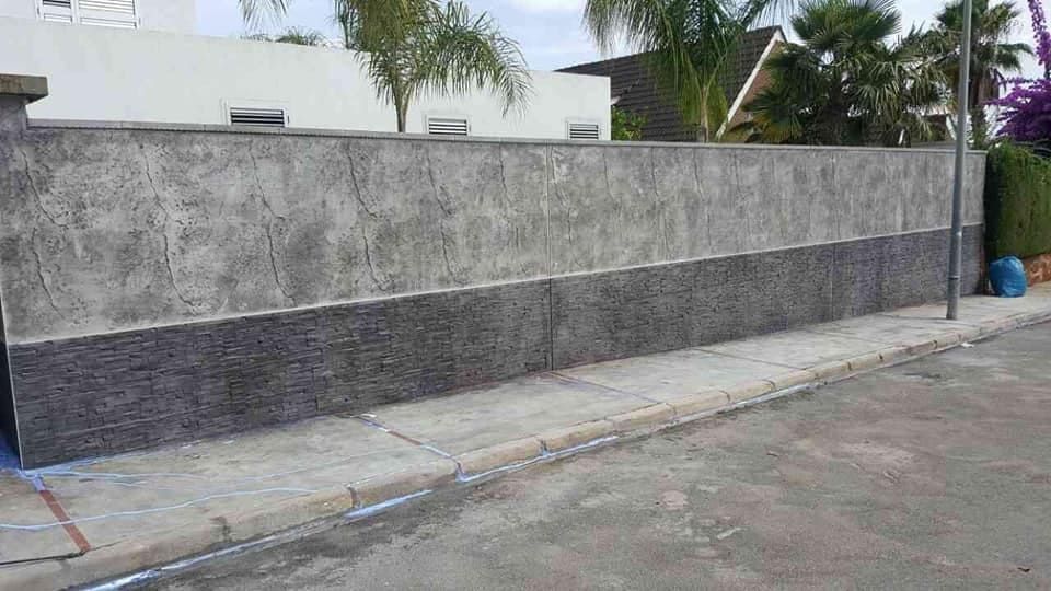 Un largo muro de hormigón bicolor junto a una acera y una calle. La parte superior es gris claro, la inferior gris oscuro.
