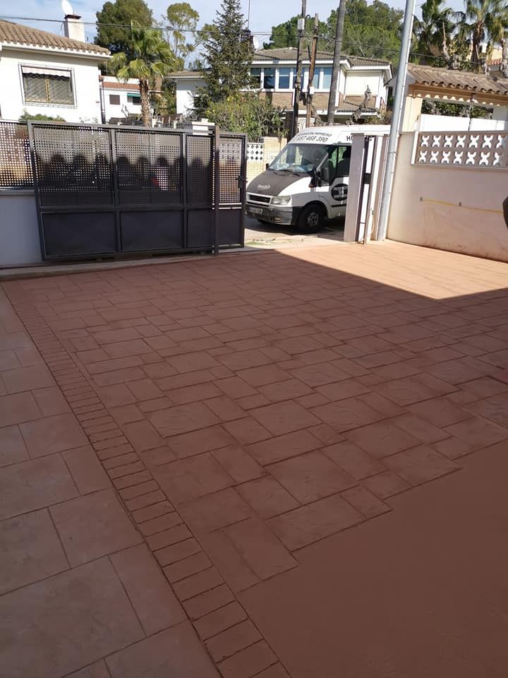 Un camino de entrada adoquinado con una puerta, una furgoneta blanca y casas al fondo.