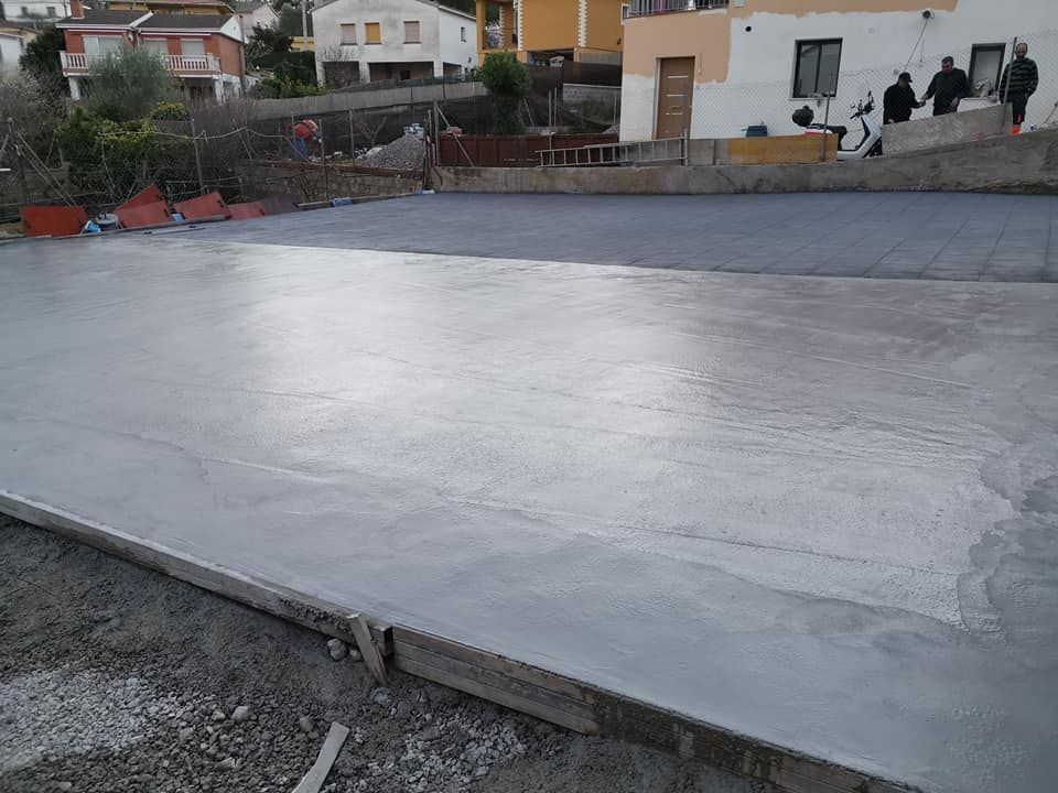 Superficie de hormigón gris recién vertido en un entorno exterior, con edificios al fondo.