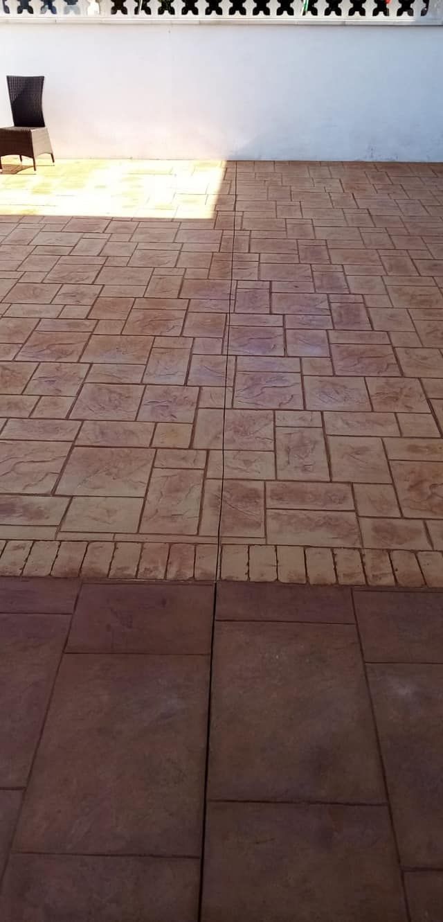 Un estrecho callejón empedrado entre un edificio de ladrillo y un muro de hormigón, que conduce hacia una zona verde.