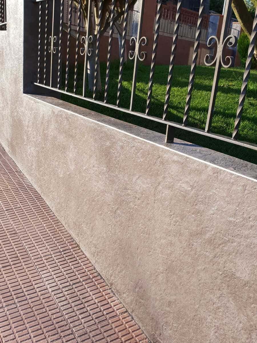 Una pared texturizada de color canela con una cerca de hierro forjado encima, un camino de ladrillos a la izquierda y un césped al fondo.