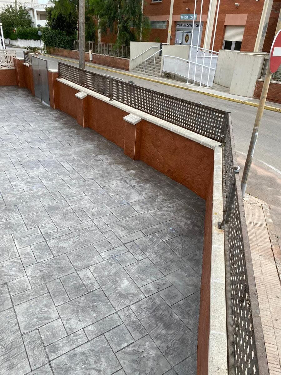 Muro de ladrillos con pavimento gris estampado y valla metálica decorativa que bordea una calle.