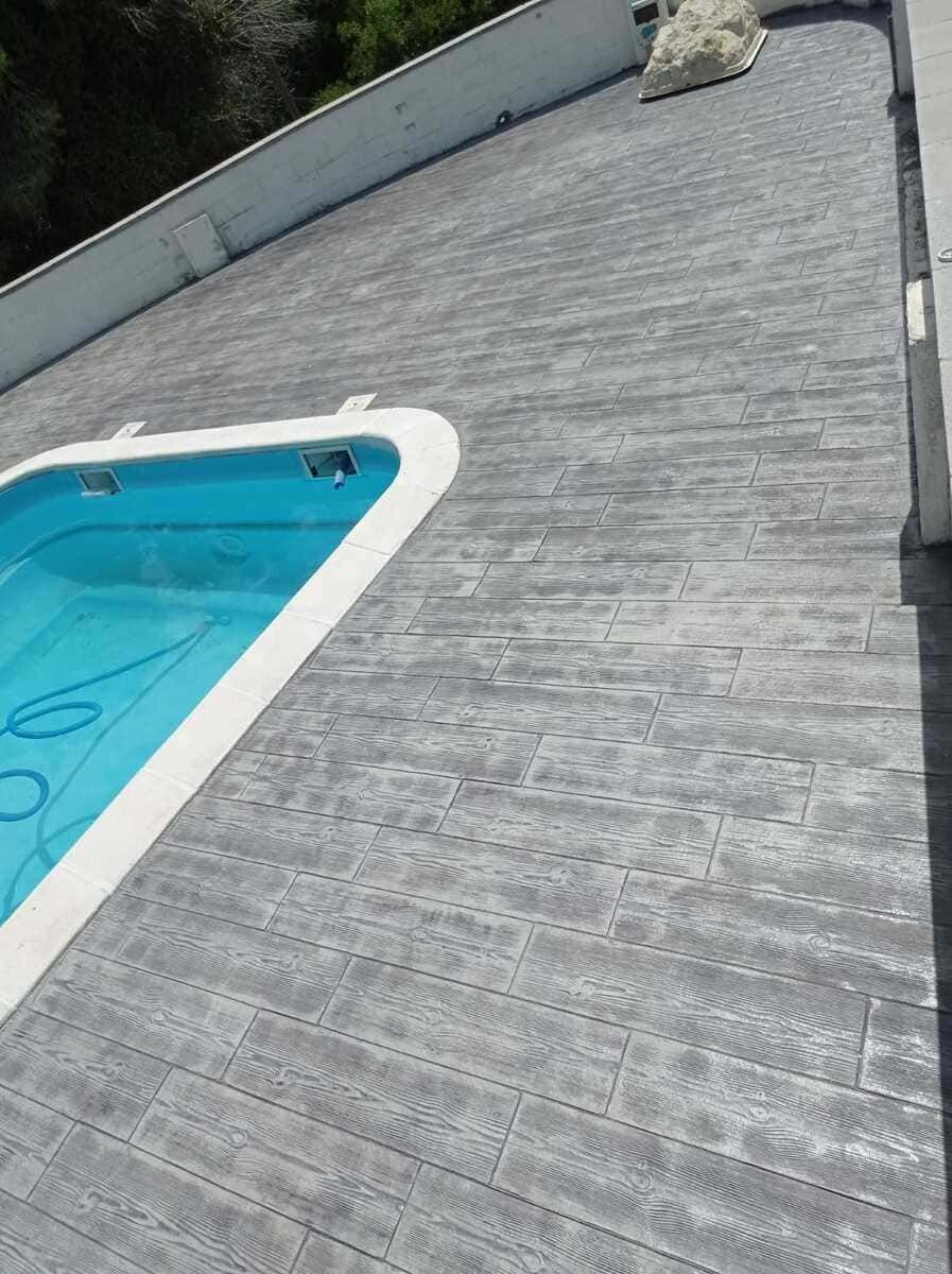 Piscina de agua azul junto a un patio de hormigón estampado gris. Borde de piscina blanco.