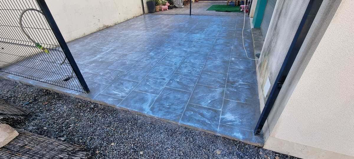 Pavimento mojado que refleja la luz, enmarcado por una pared, una valla y un borde de grava oscura.