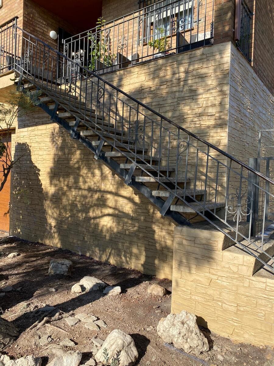 Escalera metálica exterior que conduce a la planta superior de un edificio con fachada de piedra texturizada. La luz del sol proyecta sombras.