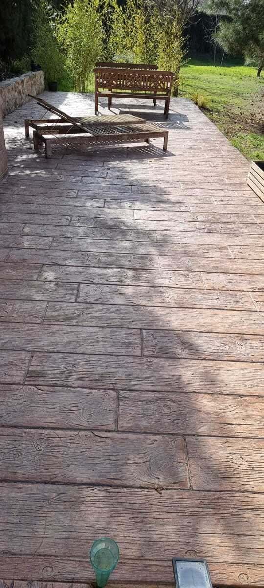Patio de madera con bancos y vegetación circundante. La luz del sol proyecta sombras sobre los tablones.