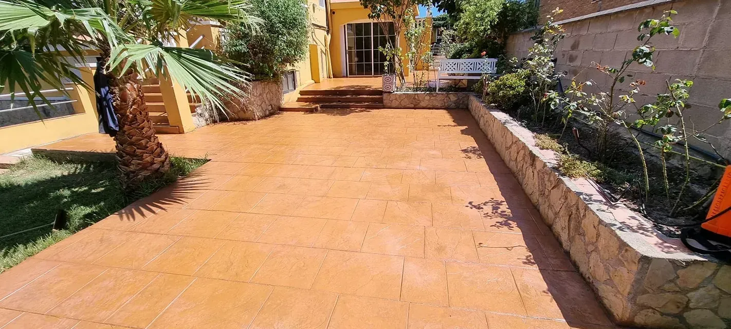 Un camino de entrada de color bronce conduce a una casa amarilla con una puerta de vidrio, enmarcada por vegetación y macizos de flores.