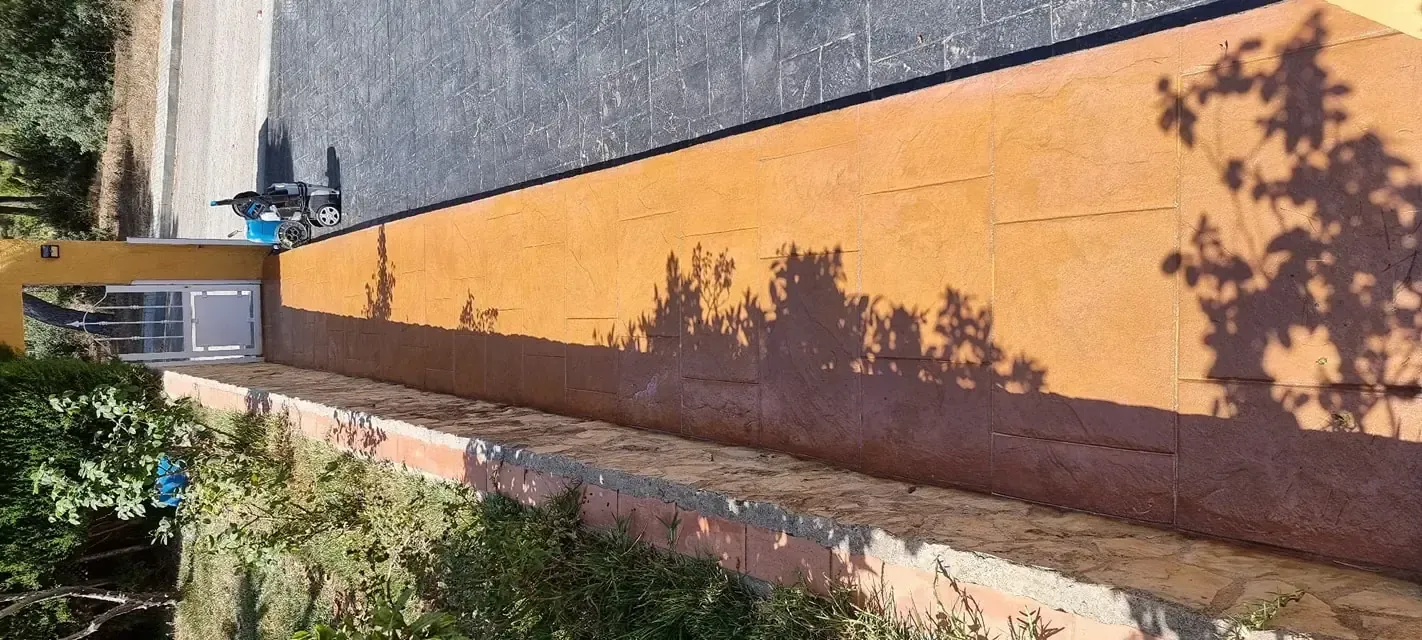 Un estrecho callejón empedrado entre un edificio de ladrillo y un muro de hormigón, que conduce hacia una zona verde.