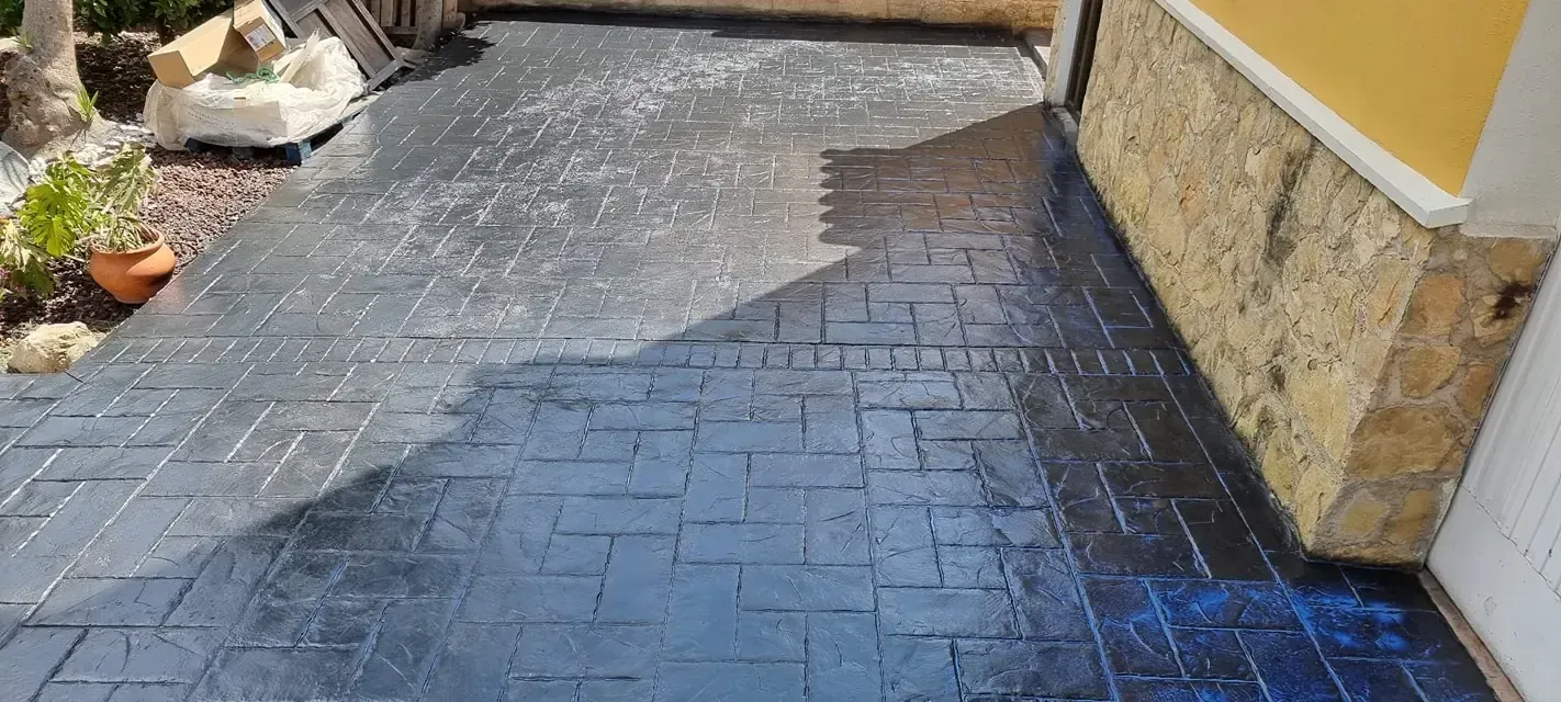 Patio de hormigón estampado gris oscuro con luz natural. Muro de piedra y parte de una casa a la derecha. Plantas y piedras a la izquierda.
