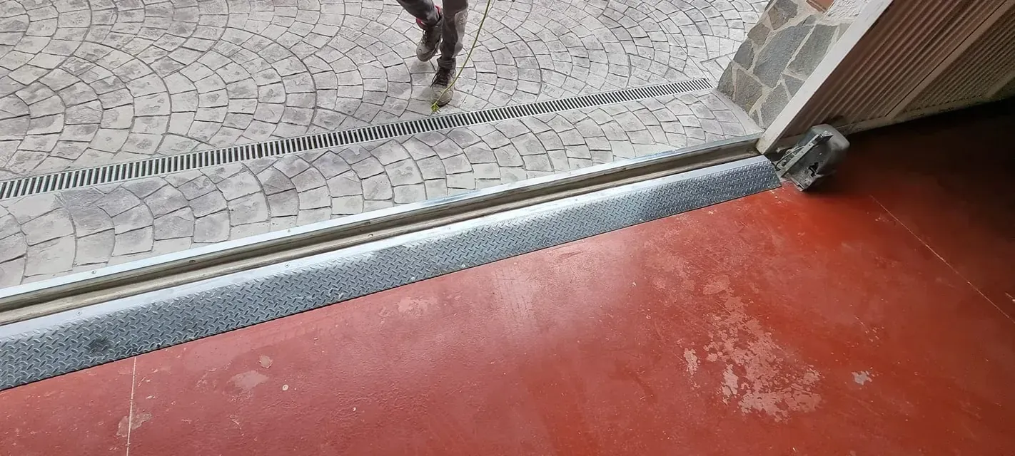 Un umbral metálico separa el suelo rojo de las baldosas grises estampadas. Se ven las piernas de una persona por encima de las baldosas.