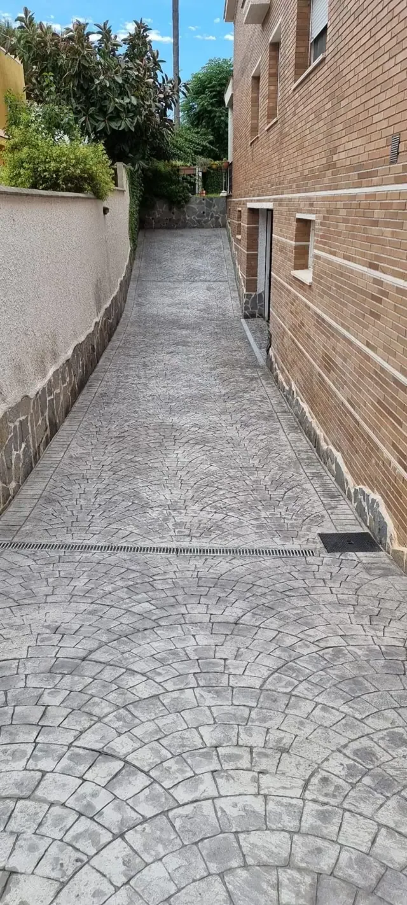 Un estrecho callejón empedrado entre un edificio de ladrillo y un muro de hormigón, que conduce hacia una zona verde.