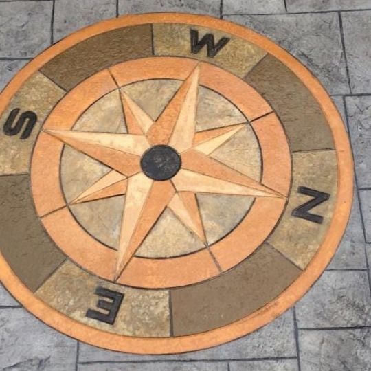 Diseño de rosa de los vientos incrustado en suelo pavimentado; tonos dorados y marrones, etiquetado con direcciones cardinales.