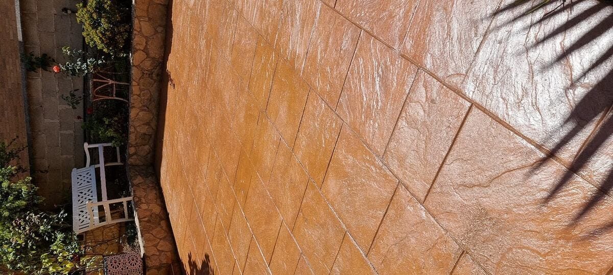Patio de hormigón con estampado marrón, un pequeño banco blanco y plantas verdes. Sombra de palmera.