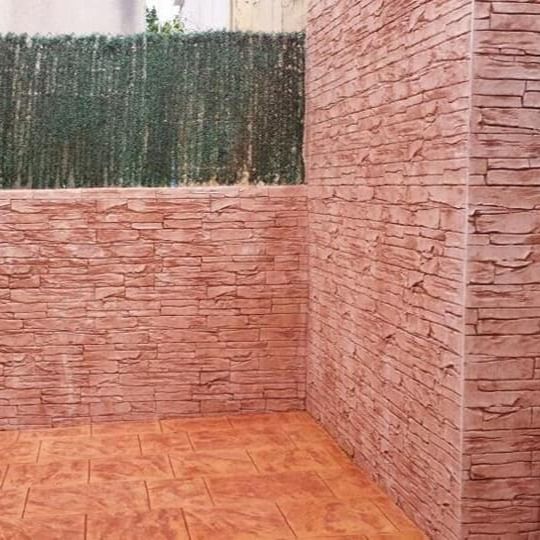 Esquina de un patio con paredes de ladrillo de piedra marrón rojiza y suelo de baldosas a juego. Al fondo, una mampara verde ofrece privacidad.